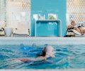 Elderly woman swimming in a modern indoor pool while another senior relaxes on a lounge chair.
