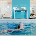 Elderly woman swimming in a modern indoor pool while another senior relaxes on a lounge chair.