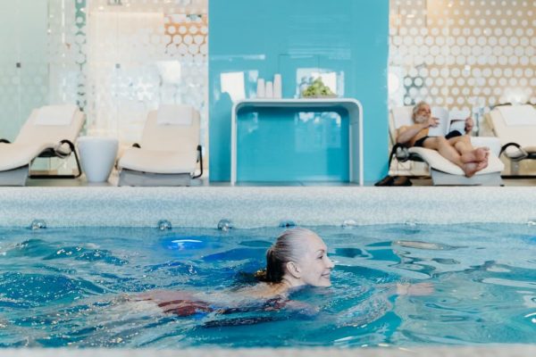 Elderly woman swimming in a modern indoor pool while another senior relaxes on a lounge chair.
