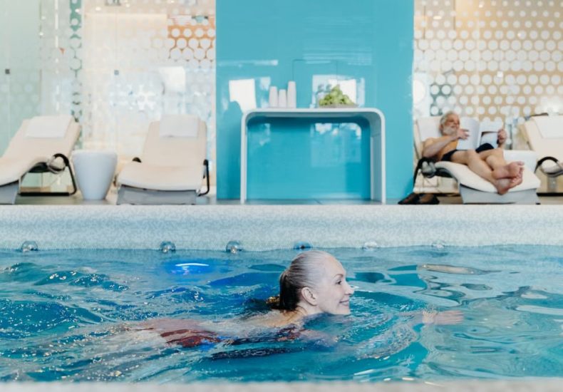 Elderly woman swimming in a modern indoor pool while another senior relaxes on a lounge chair.