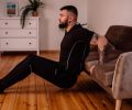 A bearded man in black fitness attire exercising at home, showcasing arm tattoos and a healthy lifestyle.